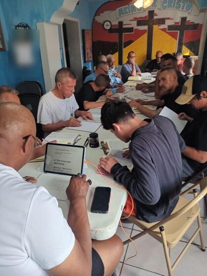 Men seated around a long table with open Bibles, beneath a mural of three crosses reading Reino de Cristo.