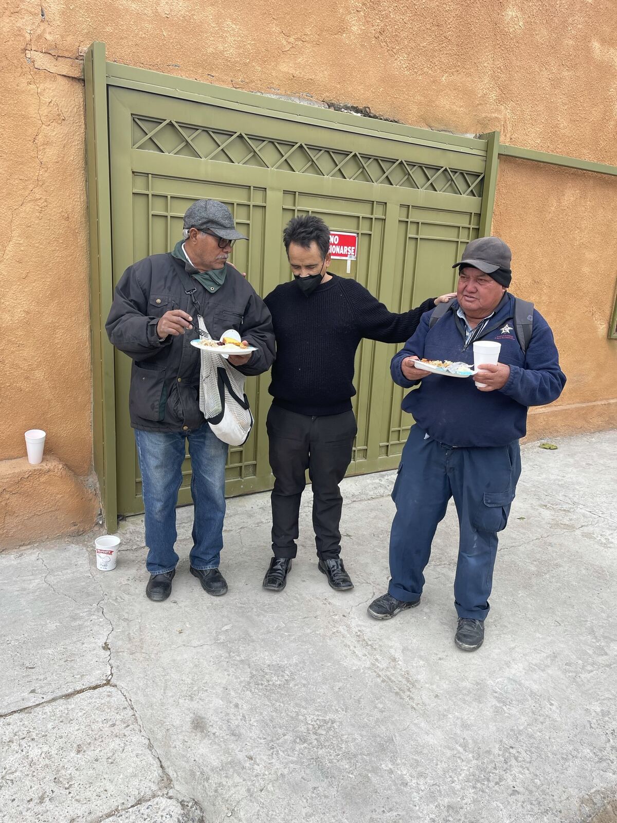Three men standing outside a green gate, two holding plates of food while the man between them puts an arm around each of them.