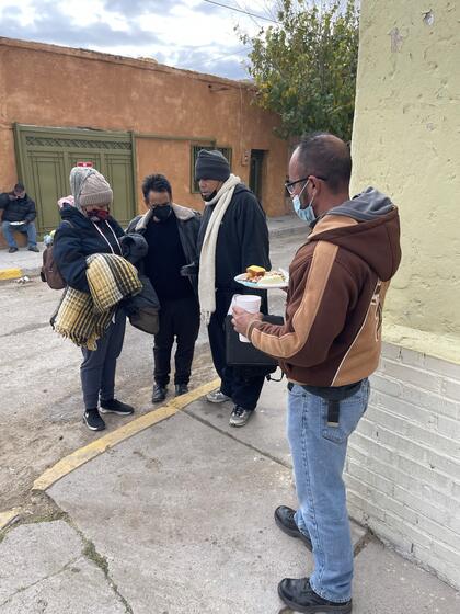 People on a street corner holding plates of food and blankets while a volunteer prays with them.