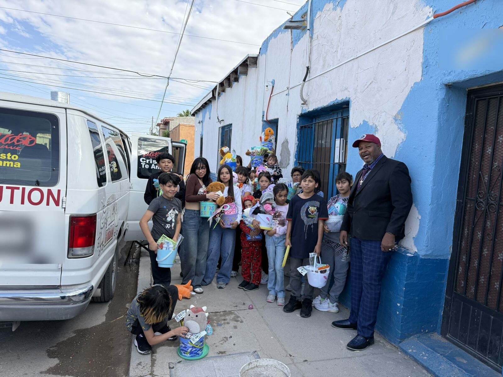 Children and young people along a street holding Easter baskets beside the ministry van, painted with the words Cristo Te Ama.
